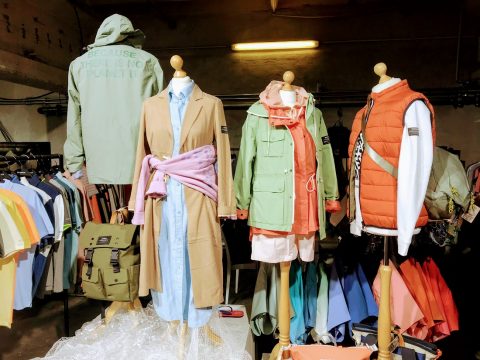 Eine Schaufensterpuppe präsentiert Outdoor-Bekleidung, darunter Jacken, Mäntel und Hemden. Die Schaufensterpuppe in der Mitte trägt einen Trenchcoat mit einem rosa Schal. Im Hintergrund hängen verschiedene bunte Hemden und Taschen an Ständern.