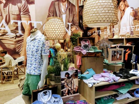 Ein Schaufenster in einem Bekleidungsgeschäft mit einer Schaufensterpuppe, die ein geblümtes Hemd und grüne Shorts trägt. Um die Schaufensterpuppe herum sind verschiedene gefaltete Hemden und Accessoires. Zwei große geflochtene Lampen hängen darüber und ein stilvoller Fotohintergrund rundet die Szene ab.