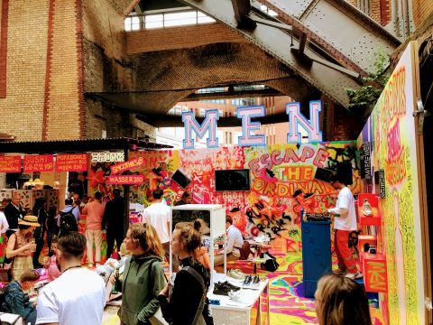 Eine lebhafte Szene mit Menschen, die sich vor einer farbenfrohen, mit Graffiti bedeckten Wand versammelt haben. Darüber sind große Buchstaben mit der Aufschrift „MEN“ zu sehen. Verschiedene Verkaufsstände und Neonschilder sind zu sehen, und Menschen stöbern und interagieren in der lebhaften Umgebung.