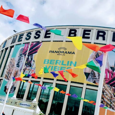 Vor dem Gebäude der Messe Berlin wehen bunte Fahnen, auf einem großen Banner steht „Panorama Berlin Vibes“. Im Hintergrund ist ein bewölkter Himmel zu sehen, darunter ein kleiner SnackPoint-Kiosk.