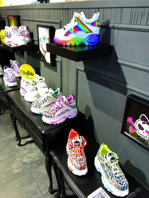 In einem Geschäft sind auf schwarzen Regalen vor einer grauen Wand farbenfrohe Buffalo London-Sneaker mit verschiedenen Animalprints und Neonfarben ausgestellt. Die Sneaker zeichnen sich durch auffällige Designs aus, darunter Leoparden- und Zebramuster.