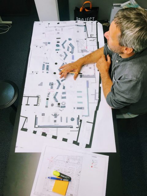 Ein Mann sitzt an einem Tisch und betrachtet große, vor ihm ausgebreitete Architekturgrundrisse. Weitere Papiere, ein Stift, ein Textmarker und Haftnotizen liegen in der Nähe. Im Hintergrund ist eine „Projekt“-Tasche zu sehen.