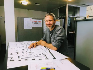 Ein Mann beugt sich über einen Schreibtisch, auf dem Architekturzeichnungen ausgebreitet sind. Er lächelt, trägt ein graues Hemd und steht in einer Büroumgebung mit einer Trennwand im Hintergrund und mehreren Dokumenten auf dem Schreibtisch.