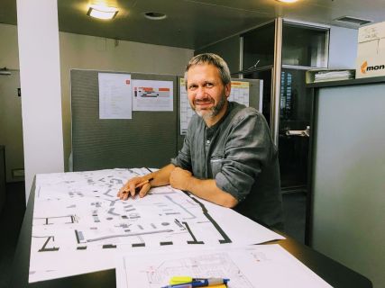 Ein Mann beugt sich über einen Schreibtisch, auf dem Architekturzeichnungen ausgebreitet sind. Er lächelt, trägt ein graues Hemd und steht in einer Büroumgebung mit einer Trennwand im Hintergrund und mehreren Dokumenten auf dem Schreibtisch.