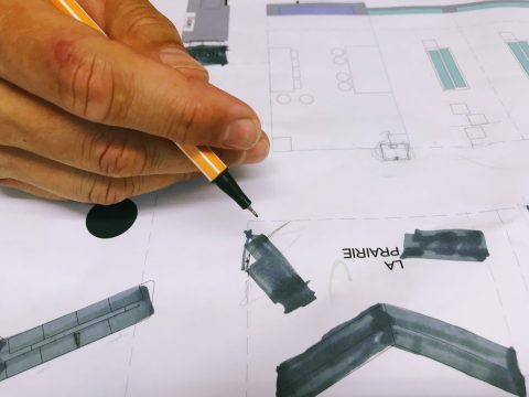 Die Hand einer Person hält einen Stift über einen Bauplan. Der Plan enthält beschriftete Abschnitte, darunter einen mit der Aufschrift „LA PRAIRIE“. Verschiedene rechteckige und kreisförmige Formen sind sichtbar, die Strukturen und Merkmale anzeigen.