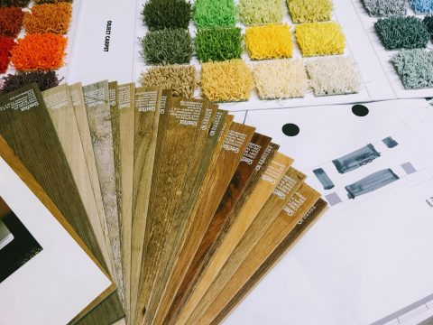 Es werden verschiedene Holz- und Teppichmuster ausgestellt. Links sind Holzbodenmuster aufgefächert und zeigen unterschiedliche Farben und Texturen. Rechts sind Teppichquadrate in verschiedenen Grün-, Gelb- und Beigetönen auf weißen Laken angeordnet.