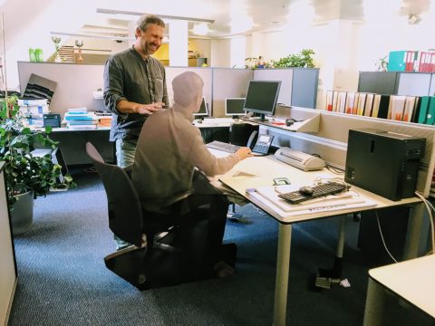 Ein Mann steht lächelnd in einem Büro, seine Hand liegt auf der Schulter einer holografischen Figur, die an einem Schreibtisch sitzt. Der Arbeitsbereich umfasst Computer, Regale mit Ordnern und verschiedene Büromaterialien.