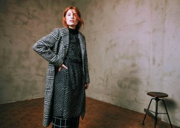 Eine Person mit roten Haaren steht selbstbewusst in einem Raum mit Betonwand. Sie trägt einen karierten Mantel über einem gepunkteten Kleid und karierten Hosen. Im Hintergrund ist ein Holzhocker auf einem Parkettboden zu sehen.