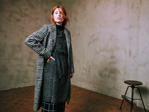 Eine Person mit roten Haaren steht selbstbewusst in einem Raum mit Betonwand. Sie trägt einen karierten Mantel über einem gepunkteten Kleid und karierten Hosen. Im Hintergrund ist ein Holzhocker auf einem Parkettboden zu sehen.