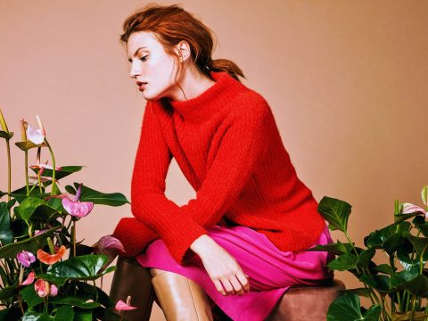 Eine Person mit roten Haaren zu einem Pferdeschwanz sitzt auf einem Hocker. Sie trägt einen roten Pullover und einen rosa Rock. Sie ist umgeben von tropischen grünen Pflanzen mit rosa und orangefarbenen Blüten vor einem schlichten Hintergrund.