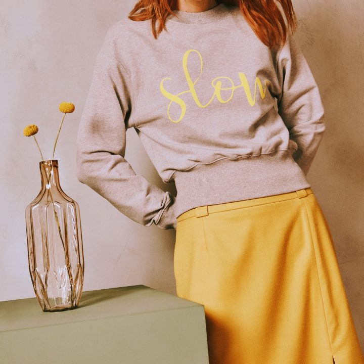 Eine Frau mit roten Haaren steht vor einer strukturierten Wand. Sie trägt ein graues Sweatshirt mit dem Wort „slow“ in Gelb und einen gelben Rock. Auf einem grünen Tisch neben ihr steht eine hohe Glasvase mit zwei gelben Blumen.