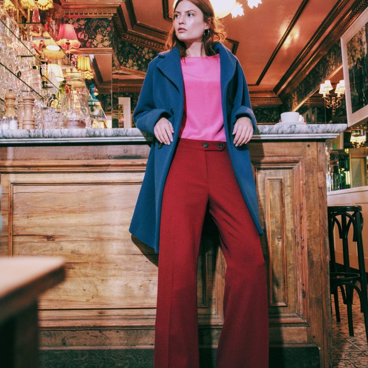 Eine Person in blauem Mantel, rosa Oberteil und roter Hose steht selbstbewusst vor einer Bar im Vintage-Stil mit Holzakzenten und gemustertem Fliesenboden. Die Atmosphäre ist mit dekorativer Beleuchtung und kunstvoller Tapete ausgestattet.