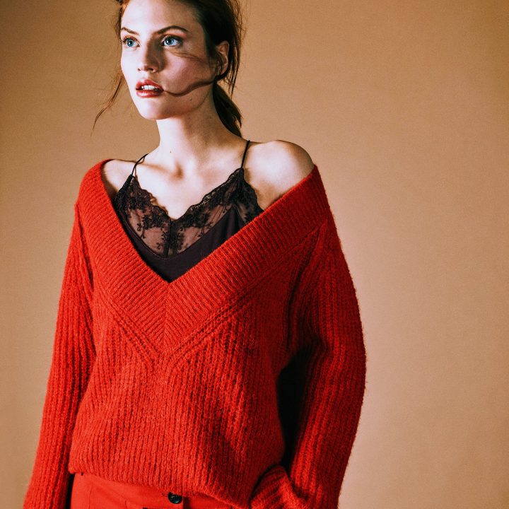 Eine Person mit langen Haaren trägt einen roten Pullover über einem schwarzen Spitzentop und roten Hosen und steht vor einem beigen Hintergrund. Ihr Pullover ist schulterfrei und sie hat einen nachdenklichen Gesichtsausdruck.