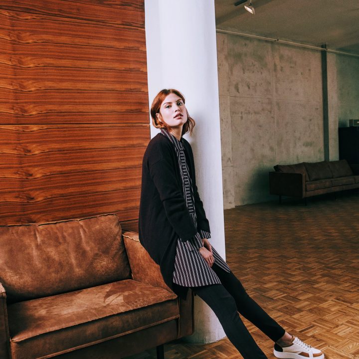 Eine Frau mit roten Haaren, die eine schwarze Strickjacke über einem gestreiften Kleid und schwarze Leggings trägt, lehnt in einem minimalistischen Raum an einer Holzwand. Sie sitzt auf einem braunen Ledersofa und trägt weiße Turnschuhe. Der Raum hat Parkettboden und Betonwände.