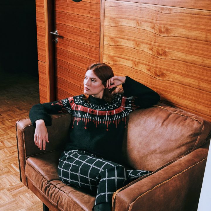 Eine Person mit braunem Haar sitzt auf einem braunen Sofa in einem holzgetäfelten Raum. Sie trägt einen schwarzen Pullover mit rot-weißem Muster, karierte Hosen und schwarze Stiefel, ein Arm ruht auf der Sofalehne.