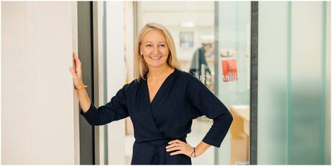 Eine Frau in einem marineblauen Kleid steht selbstbewusst da, ihre linke Hand ruht auf einer Wand und ihre rechte Hand auf ihrer Hüfte. Sie lächelt und im Hintergrund ist eine moderne Büroeinrichtung mit Glaswänden und verschwommenen Figuren zu sehen.