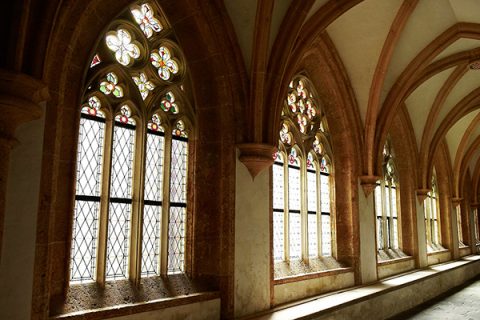 Bogenfenster im gotischen Stil füllen einen Korridor mit Licht. Die Fenster sind mit bunten Buntglasmustern geschmückt, die Spiegelbilder auf den Steinboden und die Wände werfen. Die hohen Decken werden von elegant geschnitzten Säulen getragen.