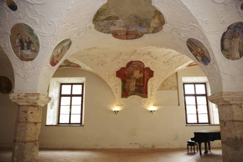 Ein historischer Raum mit kunstvollen Deckenfresken, die verschiedene Figuren darstellen. Die cremefarbenen Wände verfügen über zwei hohe Fenster und auf dem gefliesten Boden steht ein schwarzer Flügel. Das Ambiente ist klassisch und elegant.