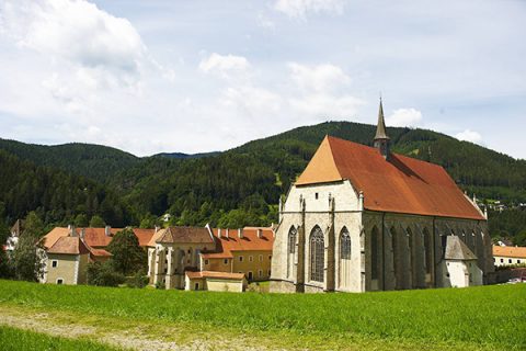 Eine mittelalterliche Abtei mit einem roten Ziegeldach steht vor einer Kulisse aus üppigen grünen Hügeln und einem teilweise bewölkten Himmel. Die umliegenden Gebäude weisen ähnliche architektonische Merkmale auf und tragen zur ruhigen Landschaft bei.