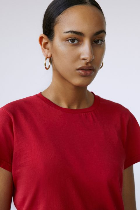 Eine Person mit zurückgekämmten dunklen Haaren trägt ein rotes T-Shirt und goldene Creolen. Der Hintergrund ist ein einfacher, heller Hintergrund.