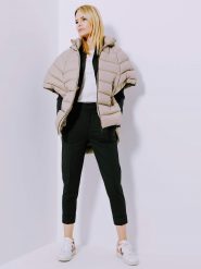 Eine Person in beigefarbener Pufferjacke, weißem T-Shirt, schwarzer Hose und weißen Turnschuhen steht mit den Händen in den Taschen vor einer weißen Wand. Sie blickt in entspannter Haltung nach oben.