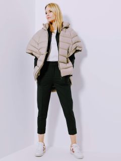 Eine Person in beigefarbener Pufferjacke, weißem T-Shirt, schwarzer Hose und weißen Turnschuhen steht mit den Händen in den Taschen vor einer weißen Wand. Sie blickt in entspannter Haltung nach oben.
