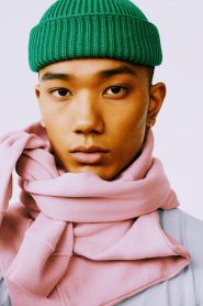 Eine Person mit einer grünen Strickmütze und einem dicken rosa Schal um den Hals blickt direkt in die Kamera. Ihr Gesichtsausdruck ist ruhig und sie steht vor einem schlichten Hintergrund.