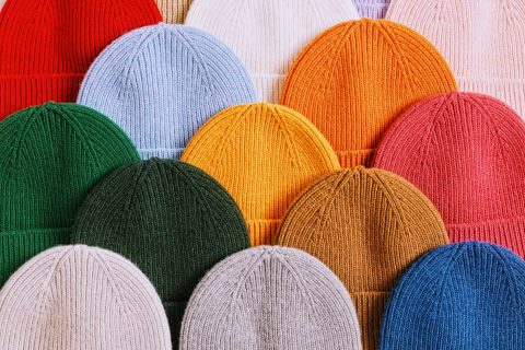 Eine Vielzahl bunter Strickmützen sind in ordentlichen Reihen angeordnet. Die Farben umfassen Rot, Rosa, Blau, Weiß, Orange, Gelb, Grün, Senfgelb und Braun und bieten eine breite Palette lebendiger und gedämpfter Töne.