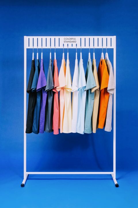 Auf einem weißen Kleiderständer hängen zwölf T-Shirts in unterschiedlichen Farben, darunter Schwarz, Lila, Blau, Rosa, Gelb, Grün, Weiß und Orange, vor einem blauen Hintergrund.