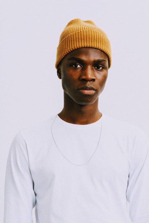 Eine Person trägt eine senffarbene Strickmütze und ein weißes Langarmshirt mit einer dünnen Halskette. Der Hintergrund ist schlicht weiß.