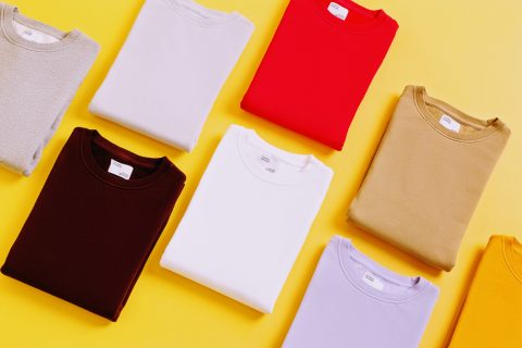 Gefaltete T-Shirts in verschiedenen Farben, darunter Rot, Weiß, Beige und Grautöne, sind ordentlich auf einem leuchtend gelben Hintergrund angeordnet.