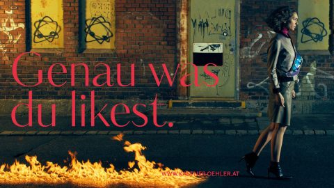 Eine Frau im Trenchcoat läuft neben einer Backsteinmauer mit Graffiti, darunter aufgesprühte Sterne. Flammen laufen neben ihr die Straße entlang. In einer großen roten Textüberlagerung steht „Genau was du likest“. Unten wird die URL einer Website angezeigt.