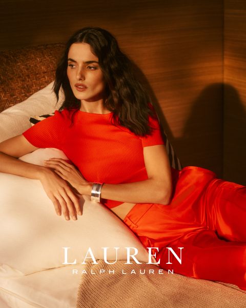 Eine Frau mit langem, gewelltem Haar lehnt auf einem weißen Sofa und trägt ein leuchtend rotes Oberteil und eine passende Hose. Warmes Sonnenlicht wirft Schatten auf die Szene. Am unteren Rand steht der Schriftzug "LAUREN RALPH LAUREN".
