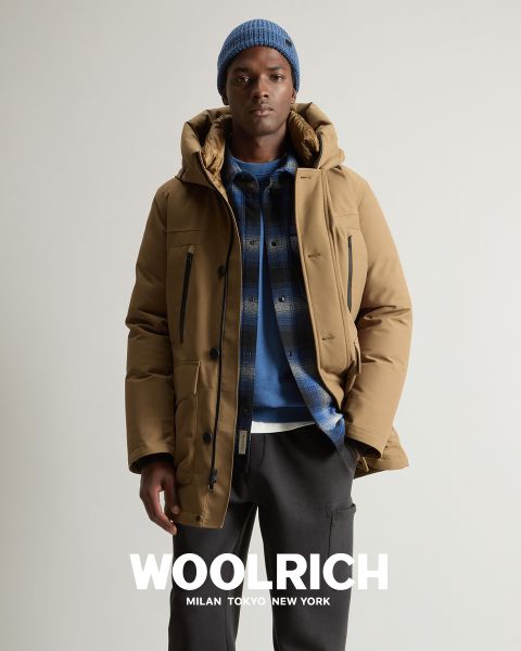 Eine Person, die einen hellbraunen Wintermantel, eine blaue Strickmütze und ein blau kariertes Hemd über einem Kapuzenpullover trägt, steht vor einem einfarbigen Hintergrund. Darunter erscheint das Wort „Woolrich“ zusammen mit den Städten Mailand, Tokio und New York.
