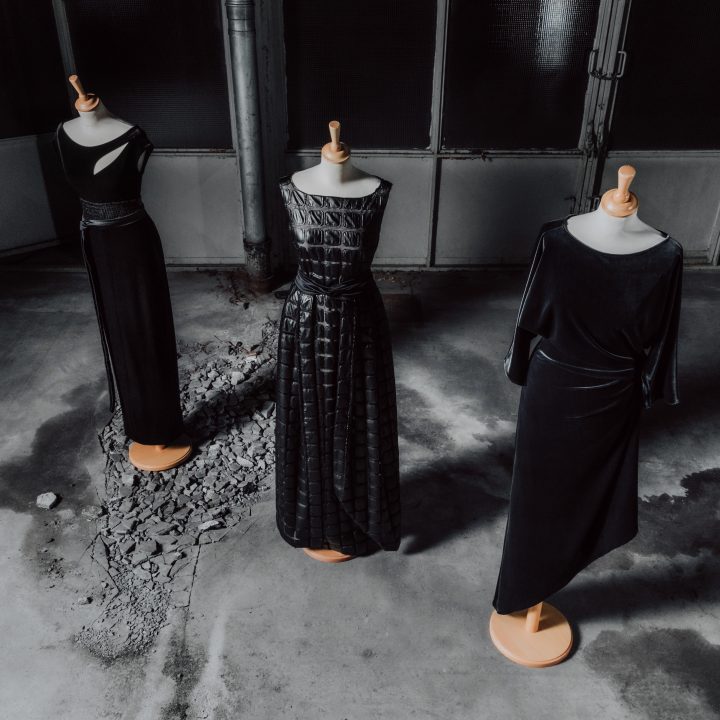 Drei Schaufensterpuppen präsentieren elegante schwarze Abendkleider. Jedes Kleid hat einen eigenen Stil und weist unterschiedliche Texturen und Schnitte auf. Sie stehen auf einem strukturierten Betonboden in einer schwach beleuchteten, industriellen Umgebung.
