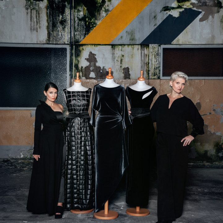 Zwei Frauen stehen neben drei Schaufensterpuppen in eleganten schwarzen Kleidern. Im Hintergrund ist eine industrielle, abgenutzte Wand mit geometrischen Mustern in Gelb und Grau zu sehen. Die Szene vermittelt eine Mischung aus moderner Mode und urbaner Ästhetik.