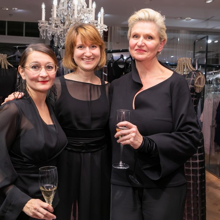 Drei Frauen in eleganten schwarzen Outfits stehen zusammen, lächeln und halten Champagnergläser in den Händen. Sie befinden sich in einem Bekleidungsgeschäft. Im Hintergrund sind Kleiderständer und ein Kronleuchter zu sehen.