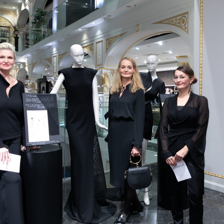 Drei schwarz gekleidete Frauen stehen neben zwei Schaufensterpuppen in eleganten schwarzen Kleidern in einem stilvollen Interieur. Sie scheinen auf einer Veranstaltung oder Ausstellung zu sein und halten Papiere in den Händen. Im Hintergrund sind dekorative goldene Akzente zu sehen.