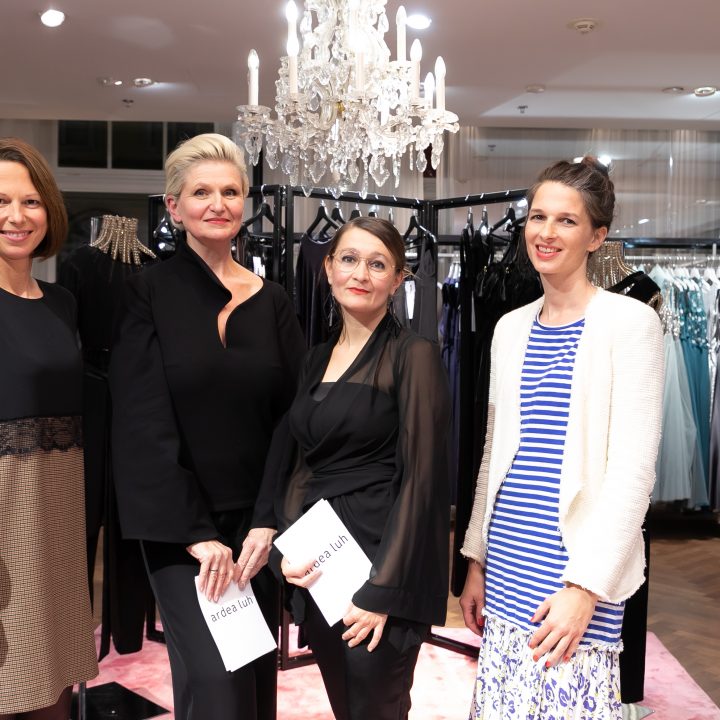 Vier Frauen stehen in einem schicken Bekleidungsgeschäft und posieren vor eleganten Kleidern, die auf Kleiderständern ausgestellt sind. Über ihnen hängt ein Kronleuchter, der der Umgebung einen Hauch von Vornehmheit verleiht. Zwei Frauen halten Veranstaltungsbroschüren in den Händen.