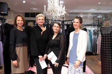 Vier Frauen stehen vor einer Kleiderausstellung mit eleganten Kleidern. Über ihnen hängt ein Kronleuchter. Sie halten Veranstaltungsbroschüren in den Händen und sind in stilvolle, halbformelle Kleidung gekleidet. Die Kulisse scheint eine Boutique oder ein Ausstellungsraum zu sein.