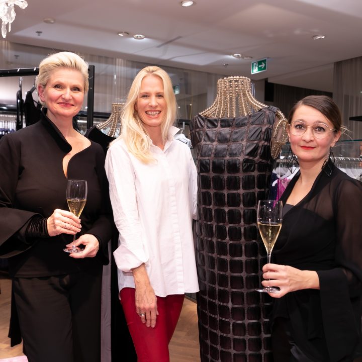 Drei Frauen stehen lächelnd mit Champagnergläsern in den Händen in einem Bekleidungsgeschäft. Neben ihnen steht eine Schaufensterpuppe in einem gemusterten Kleid. Im Hintergrund sind Kleiderständer und ein Kronleuchter über dem Kopf zu sehen.
