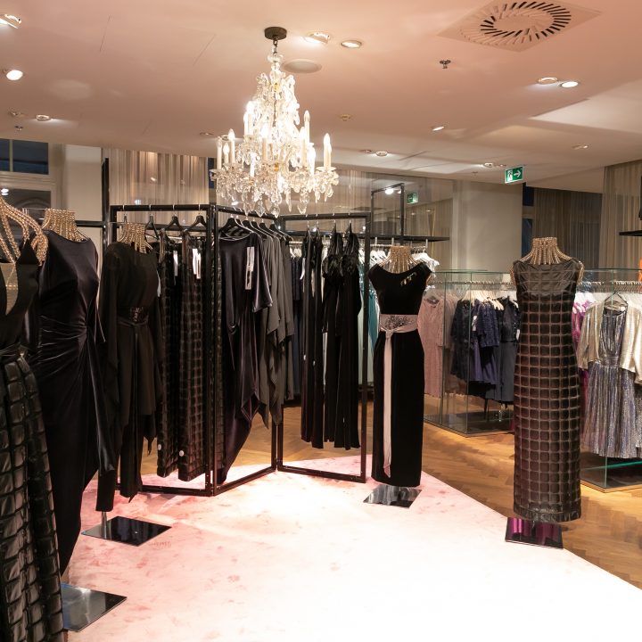 Inneneinrichtung eines Boutique-Geschäfts mit eleganten schwarzen Kleidern auf Schaufensterpuppen, die von einem Kronleuchter beleuchtet werden. Der Holzboden und die Glasregale im Hintergrund zeigen verschiedene Kleidungsstücke und schaffen so eine luxuriöse Einkaufsatmosphäre.