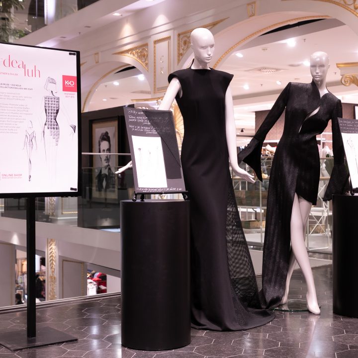 In einem luxuriösen Geschäft sind zwei Schaufensterpuppen in eleganten schwarzen Kleidern ausgestellt. Die Kulisse umfasst kunstvolles Dekor und Informationstafeln zu den Outfits.