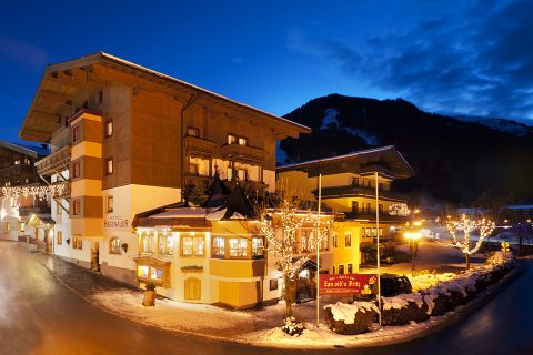 Eine malerische Dorfszene in der Abenddämmerung zeigt ein warm erleuchtetes Hotel, das mit festlichen Lichtern geschmückt ist. Schnee bedeckt den Boden und Berge heben sich als Silhouetten vor einem tiefblauen Himmel ab. Die Straßen sind ruhig und von orangefarbenen Straßenlaternen beleuchtet.