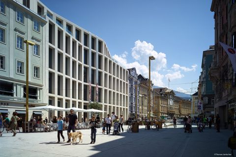 Eine belebte Fußgängerzone mit Menschen, die in Straßencafés spazieren gehen und essen. Moderne und historische Gebäude säumen die Straße unter einem klaren blauen Himmel mit vereinzelten Wolken. Ein Golden Retriever läuft neben einer Person her und im Hintergrund ist eine Bergkette zu sehen.