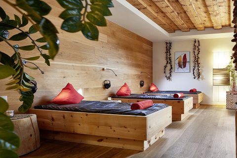 Ein gemütlicher Spa-Raum verfügt über drei Holzliegen mit roten Kissen und gerollten Handtüchern. Warmes Licht und Holzwände sorgen für eine entspannende Atmosphäre. Im Vordergrund hängen grüne Pflanzen und verleihen einen Hauch von Natur.