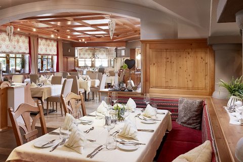 Ein gemütliches Restaurant-Interieur mit Holzpaneelen und -decke. Die Tische sind mit weißen Tischdecken, elegantem Geschirr und Servietten gedeckt. Natürliches Licht fällt durch die Fenster und verstärkt die warme und einladende Atmosphäre.