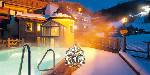 Eine gemütliche Winterszene zeigt einen beheizten Außenpool, der von Schnee umgeben ist. Warme Lichter beleuchten das nahe gelegene Gebäude und schaffen eine einladende Atmosphäre. Im Hintergrund ist eine Berglandschaft unter einem klaren Abendhimmel zu sehen.