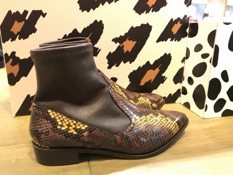 Ein Paar stylische Stiefeletten mit Schlangenhautmuster und dunklem Lederbesatz vor dem Hintergrund abstrakter Animalprint-Designs.