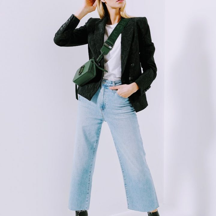 Eine Person steht selbstbewusst in einem minimalistischen Ambiente, trägt einen schwarzen Blazer über einem weißen Hemd, hellblaue Cropped-Jeans und schwarze Stiefeletten. Sie hat eine grüne Umhängetasche an und macht eine stylische Pose.